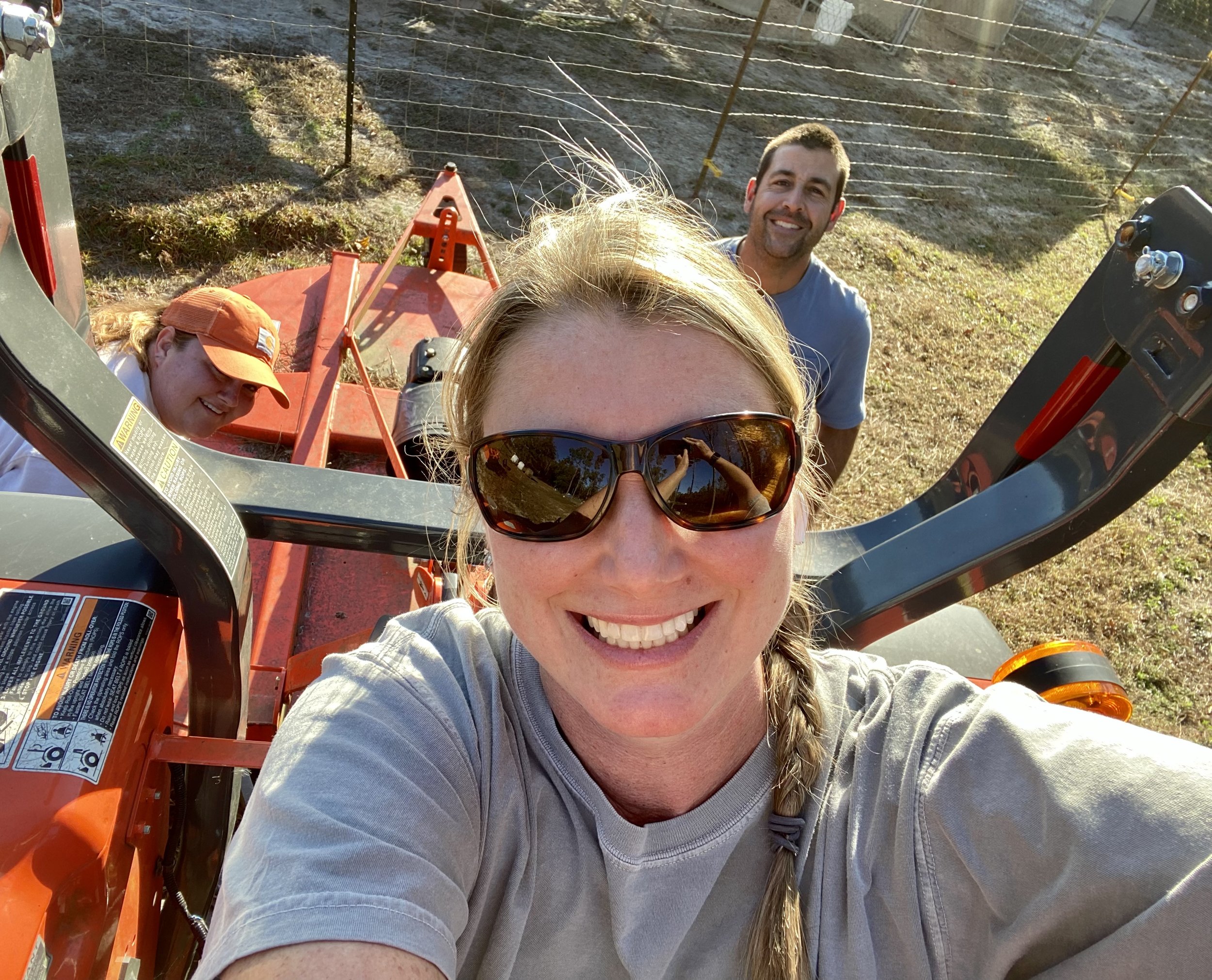 Farmasea+North+Carolina+-+2C8A5B6C-7C42-4DB3-AFCD-820F69A685E5 Three people smiling in a selfie on a tractor, with a sunny outdoor backdrop and a fence in the background, showcasing teamwork in agricultural activities.