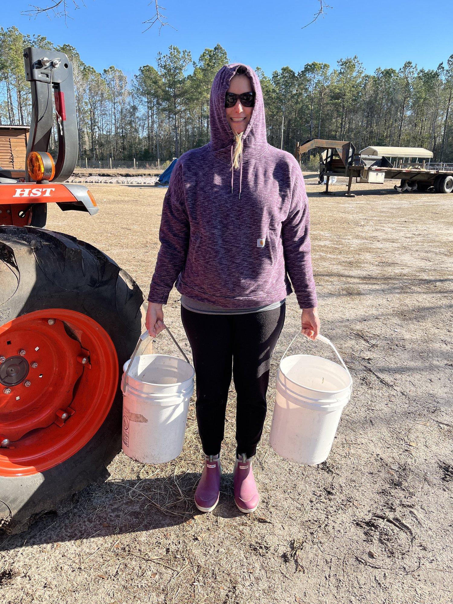 Farmasea+North+Carolina+-+89A9E728-DB56-4594-A3D6-D29FFDDCC710 Woman wearing a purple hoodie and sunglasses stands outdoors next to a tractor, carrying two white buckets in her hands. The background features trees and equipment, indicating a rural or farm setting.