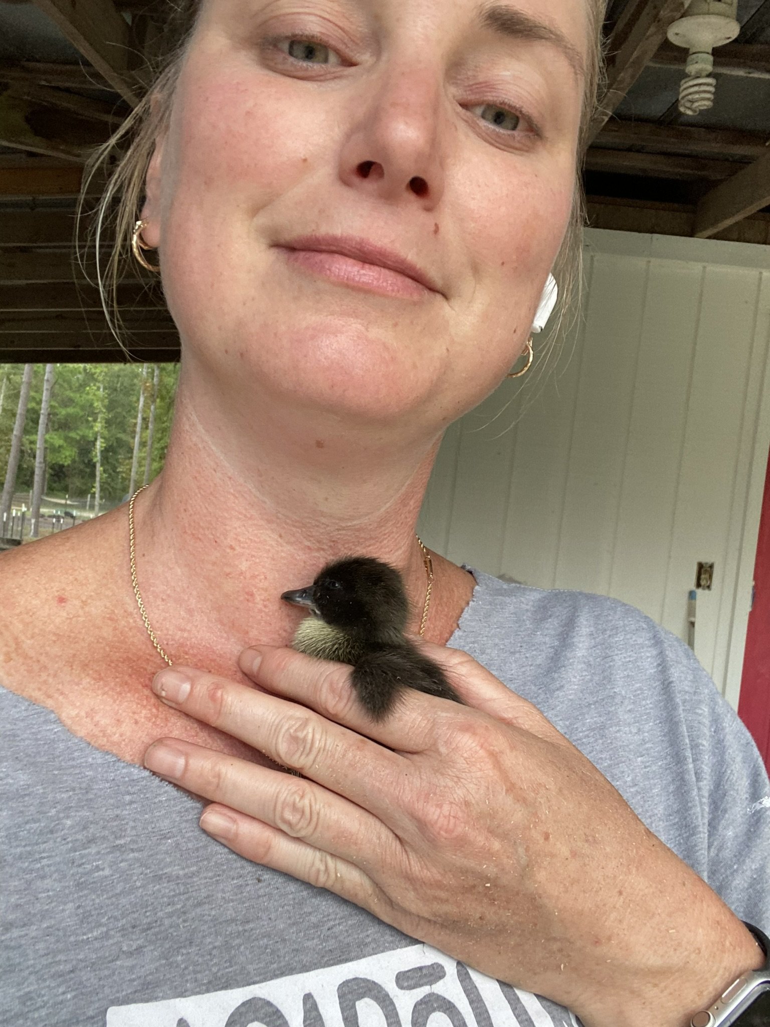 Farmasea+North+Carolina+-+8DC9AC04-C2D3-40F6-A71F-B38EF4B04D99 Woman holding a small duckling close to her neck, smiling gently, with a rustic background. The image captures a warm moment of connection between the person and the animal, highlighting themes of compassion and care for wildlife.