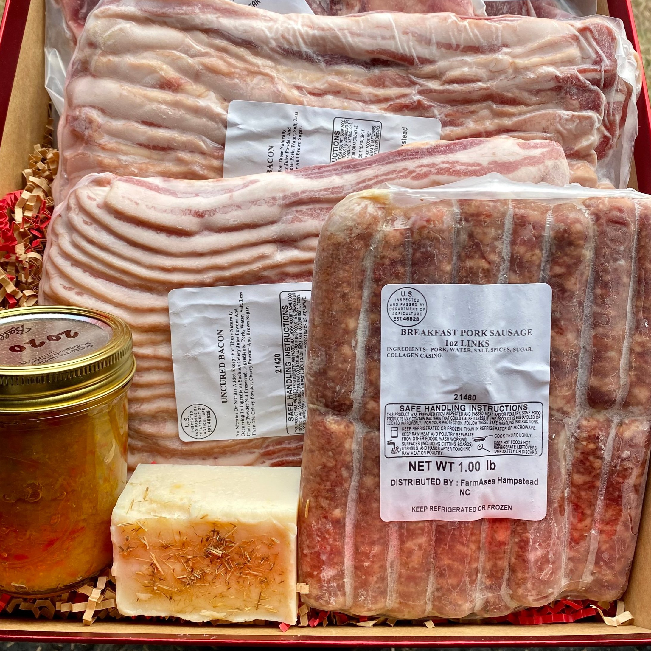 Assorted gourmet meats including uncured bacon, breakfast pork sausage links, and a jar of homemade preserves, packaged in a gift box with straw filler. Ideal for breakfast enthusiasts and food lovers.