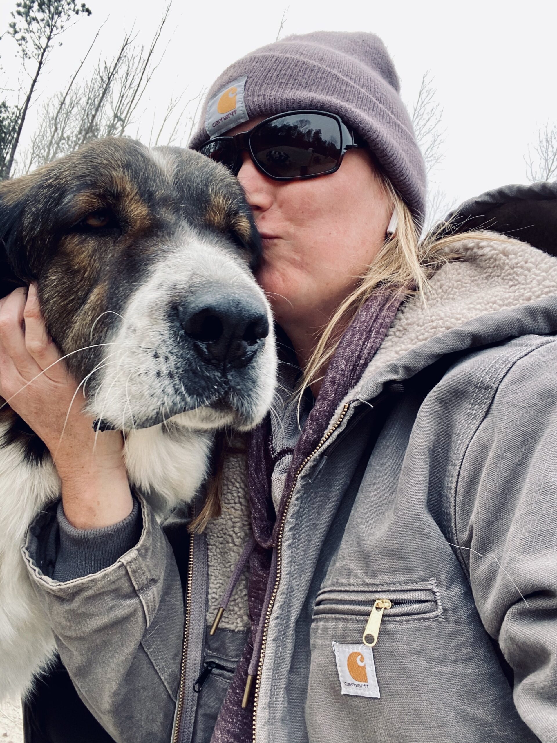 Woman wearing a beanie and sunglasses kisses a large dog on the cheek, showcasing a moment of affection in an outdoor setting with bare trees in the background.