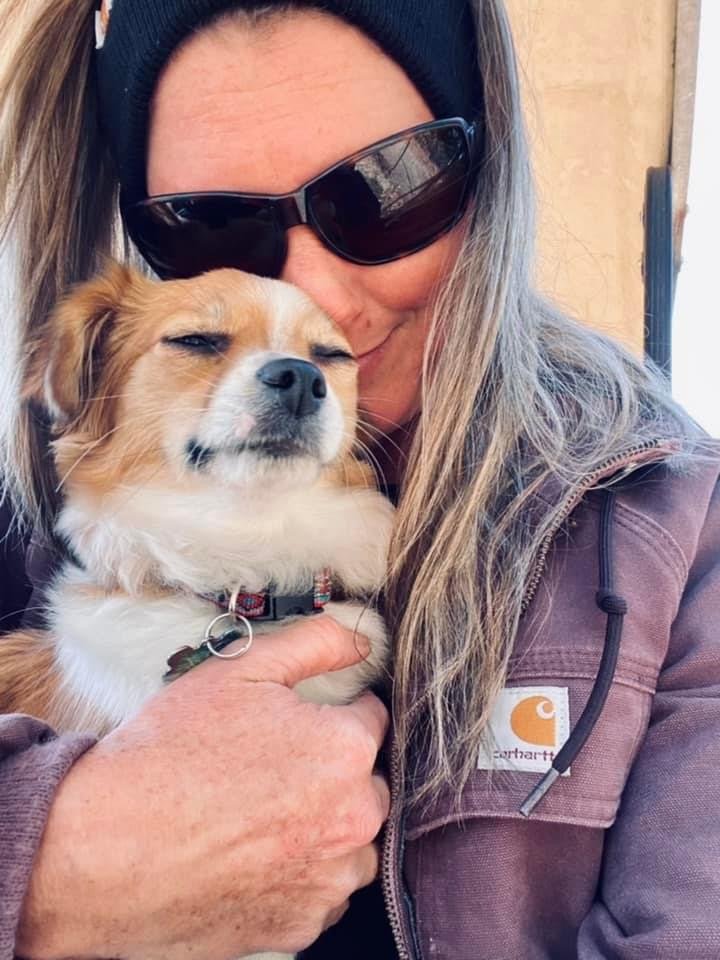 Farmasea+North+Carolina+-+F52440B2-A962-4CA2-93E6-077756BBE616 A woman wearing sunglasses and a beanie smiles while holding a small dog close to her face, showcasing a joyful bond between them. The woman is dressed in a brown jacket, and the setting appears to be outdoors.