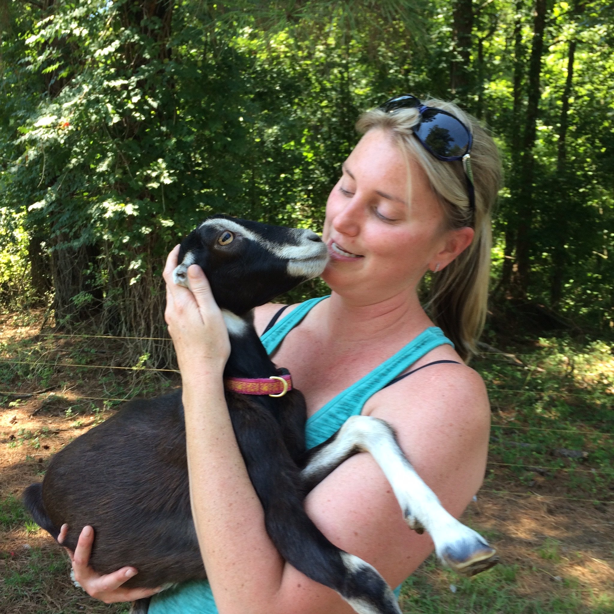 IMG_0028 Woman holding a black goat while smiling in a lush, green outdoor setting, showcasing a bond between humans and animals.