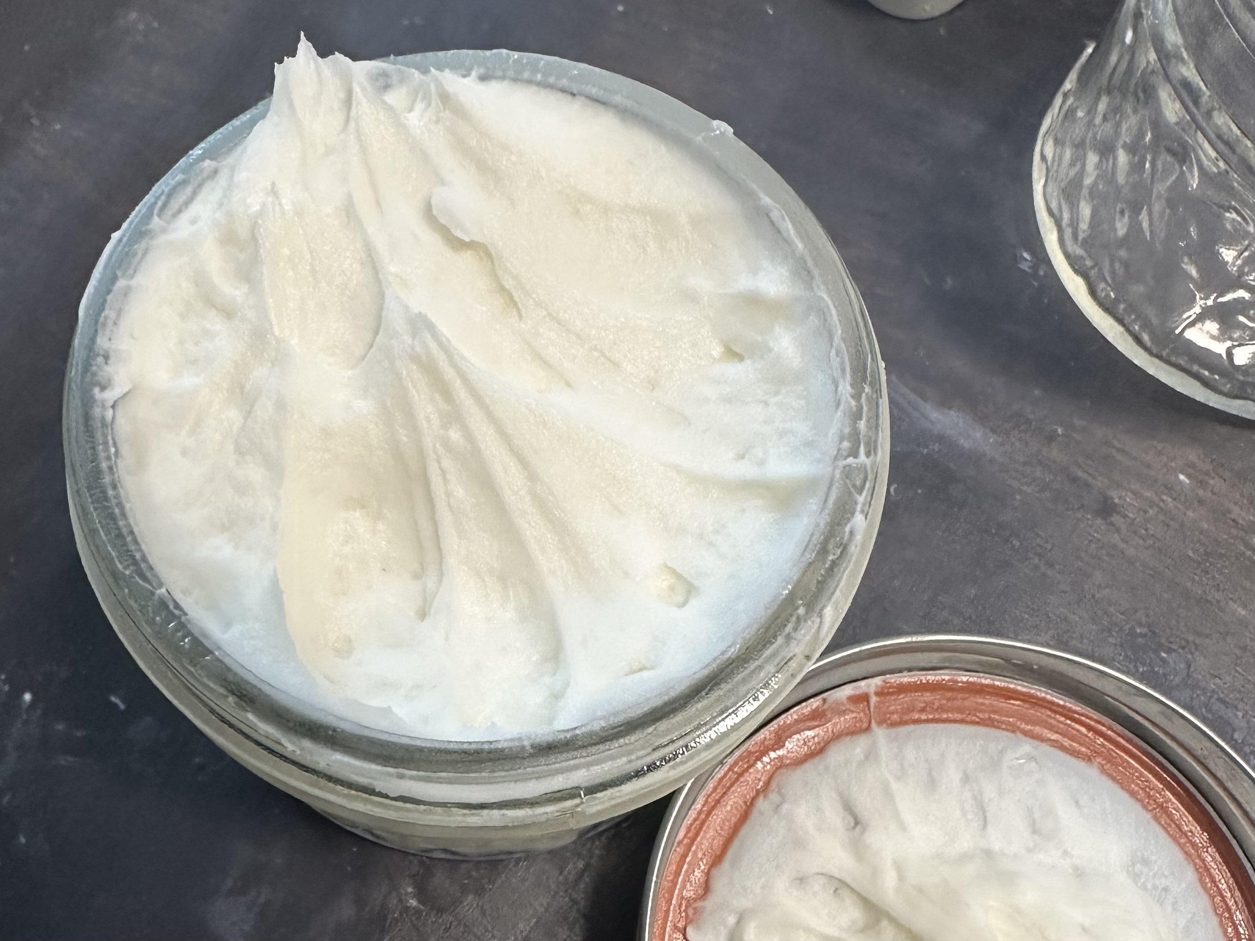 Close-up of a jar filled with creamy, whipped body butter, showcasing a smooth and fluffy texture. The jar is placed on a dark surface, highlighting the product's rich consistency and luxurious appearance.